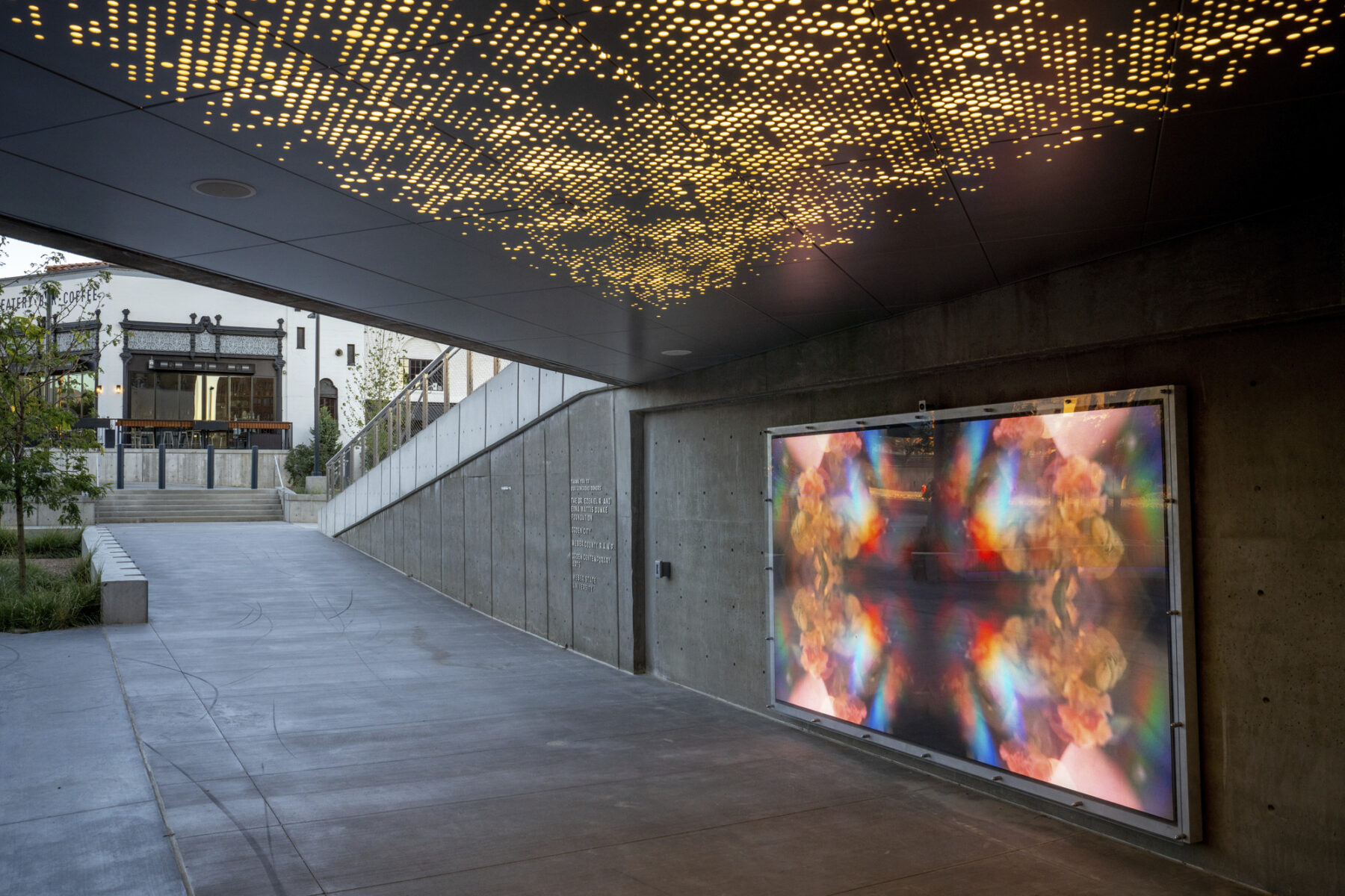 perspective photograph of art displayed on wall beneath plaza overlook with perforated ceiling and light shining through