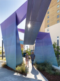 photograph of sculptural beacon with sunlight glaring through the perforations and a couple walking through pavilion