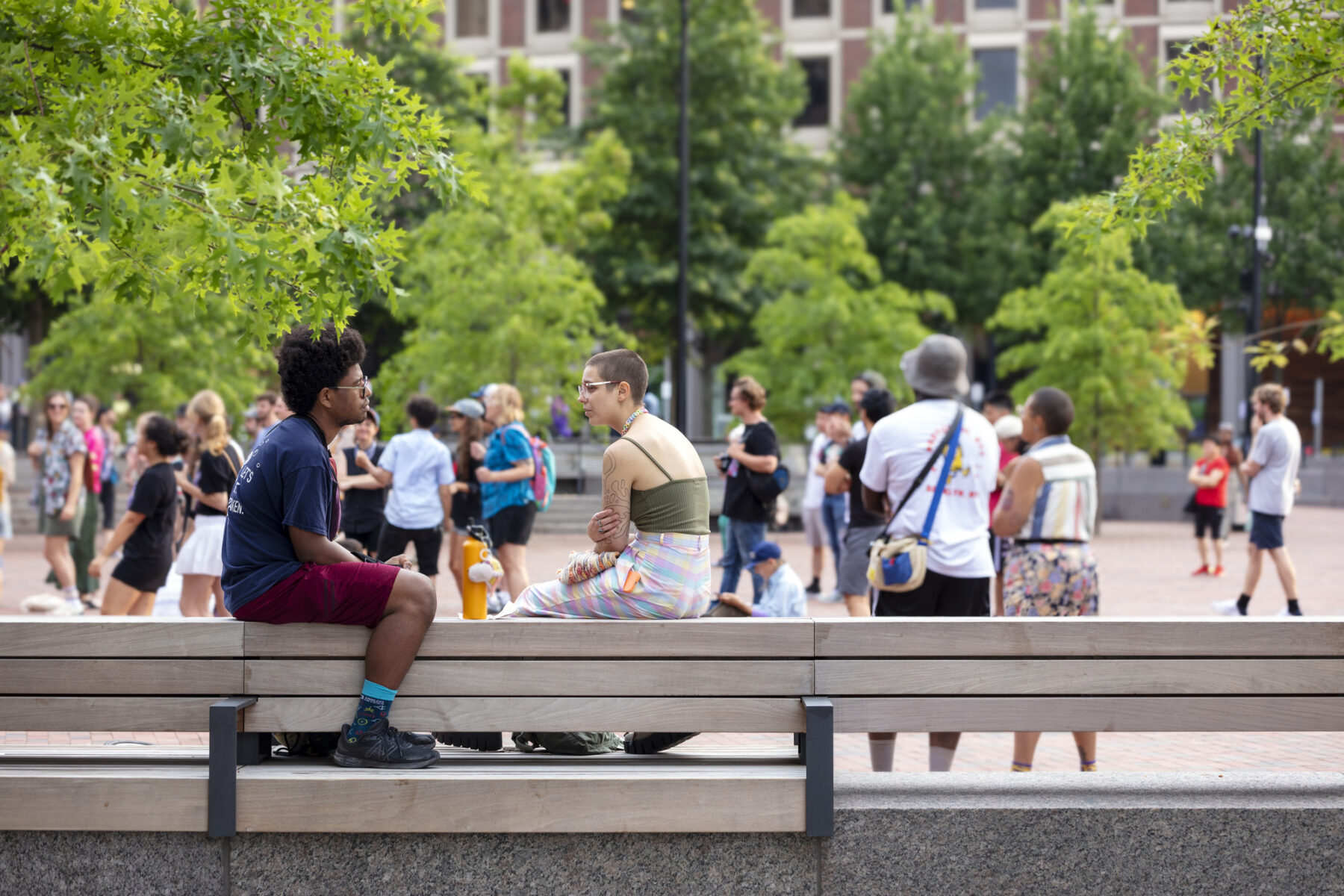 Photo of two people sitting on new plaza bench with tree canopy and public programmed event in the background