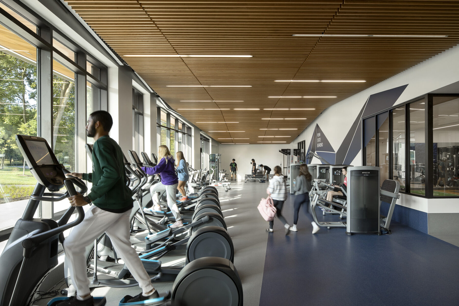 interior photograph of lively fitness area, treadmills and ellipticals on the left, weight machines on the right. Wood slatted ceiling with linear lights